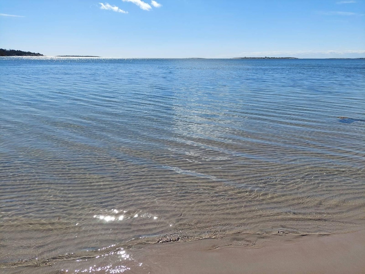 Picture from a hidden gem of a beach with crystal clear water and little ripples froming in the sand with blue sky, and some small islands in the distance.