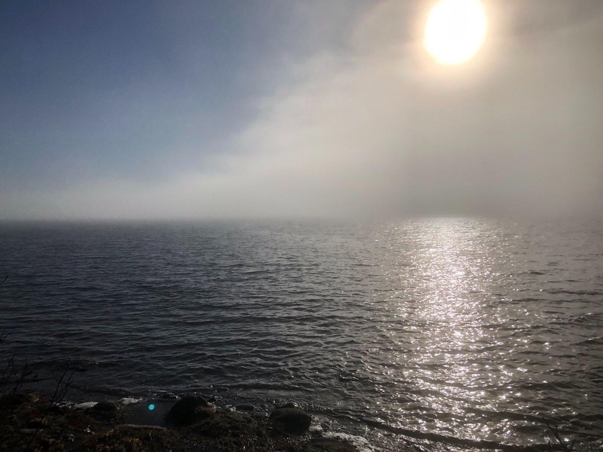 Photo of the sea from shore with heavy fog partially hiding the sun on the right and blue skies on the left.
