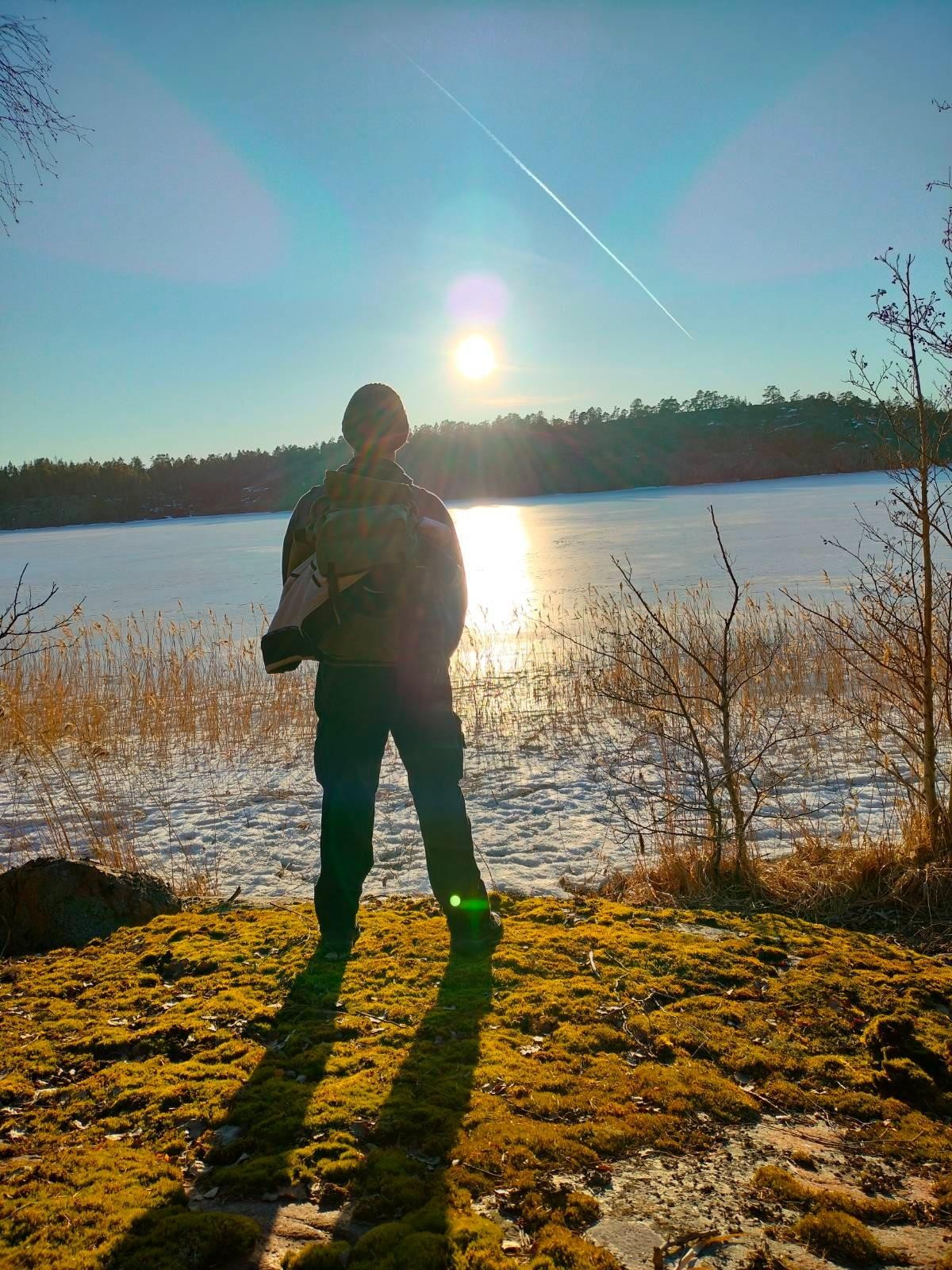 Photo of my back standing by the shore enjoying the clear blue sky and the sun shining in over the treeline in the other side of the frozen bay.