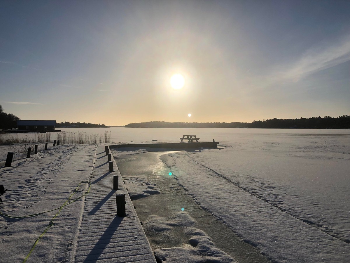Photo of a snowy bay from a bridge with a table at the end. In the distance forest covered islands. Above a blue sky and a shinning sun.
